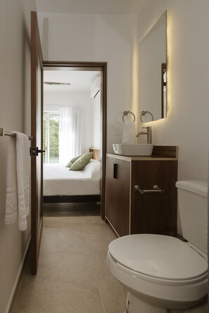 Un baño moderno con un lavamanos blanco, un inodoro y una cama visible a través de una puerta abierta.