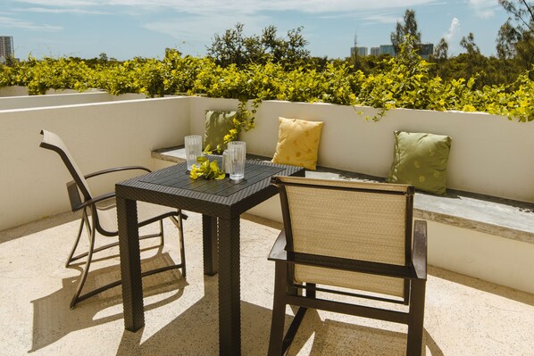 Una terraza en la azotea con una mesa y sillas, un sofá con cojines, y vistas al horizonte urbano y zonas verdes.