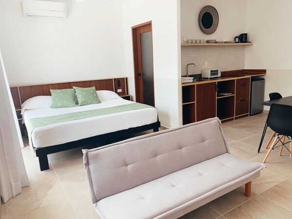 Un dormitorio con cama, mesita de noche, banco, kitchenette y puerta de baño.