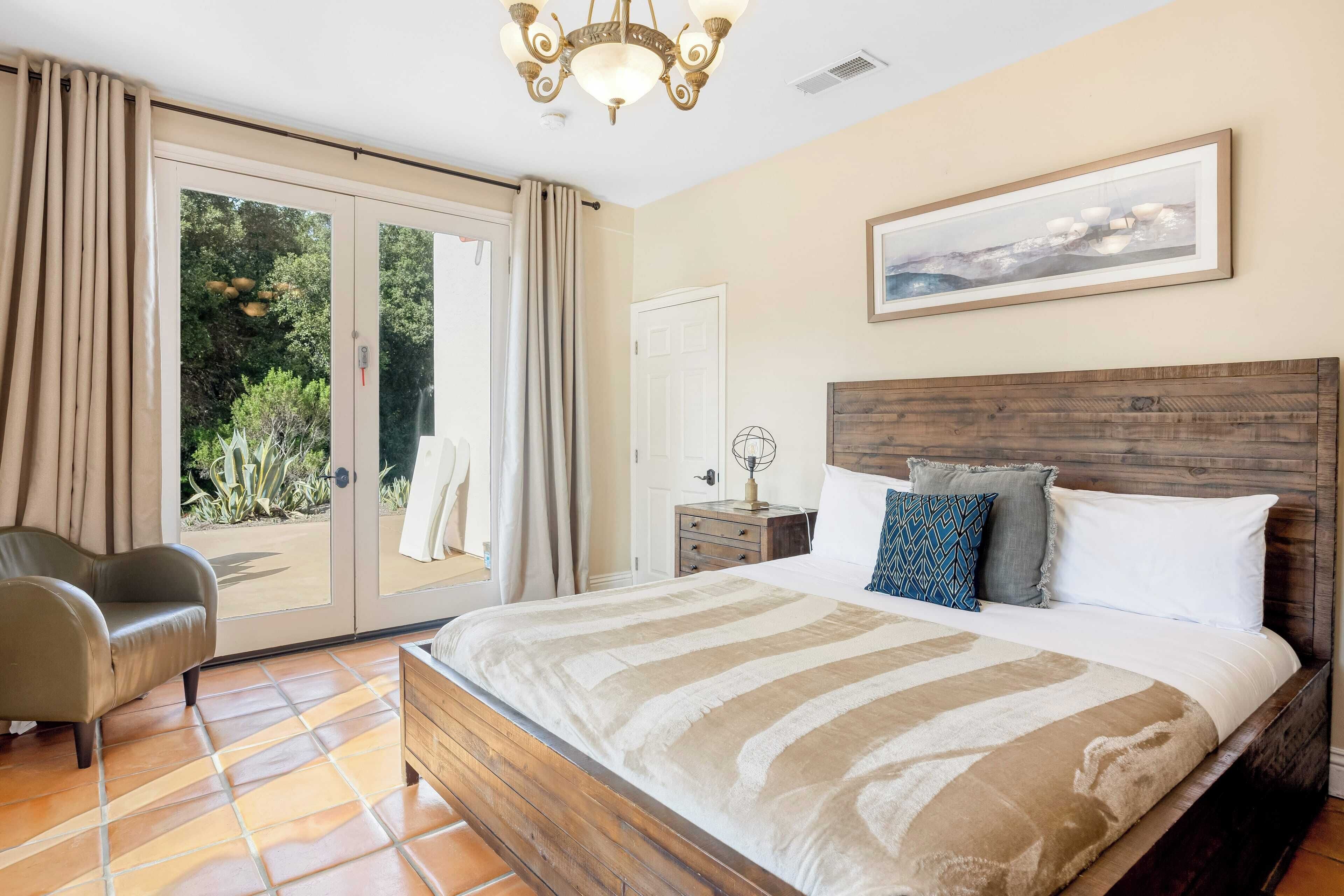 Une chambre avec un grand lit, une tête de lit en bois, une table de chevet, une chaise et une porte-fenêtre coulissante donnant sur un jardin.