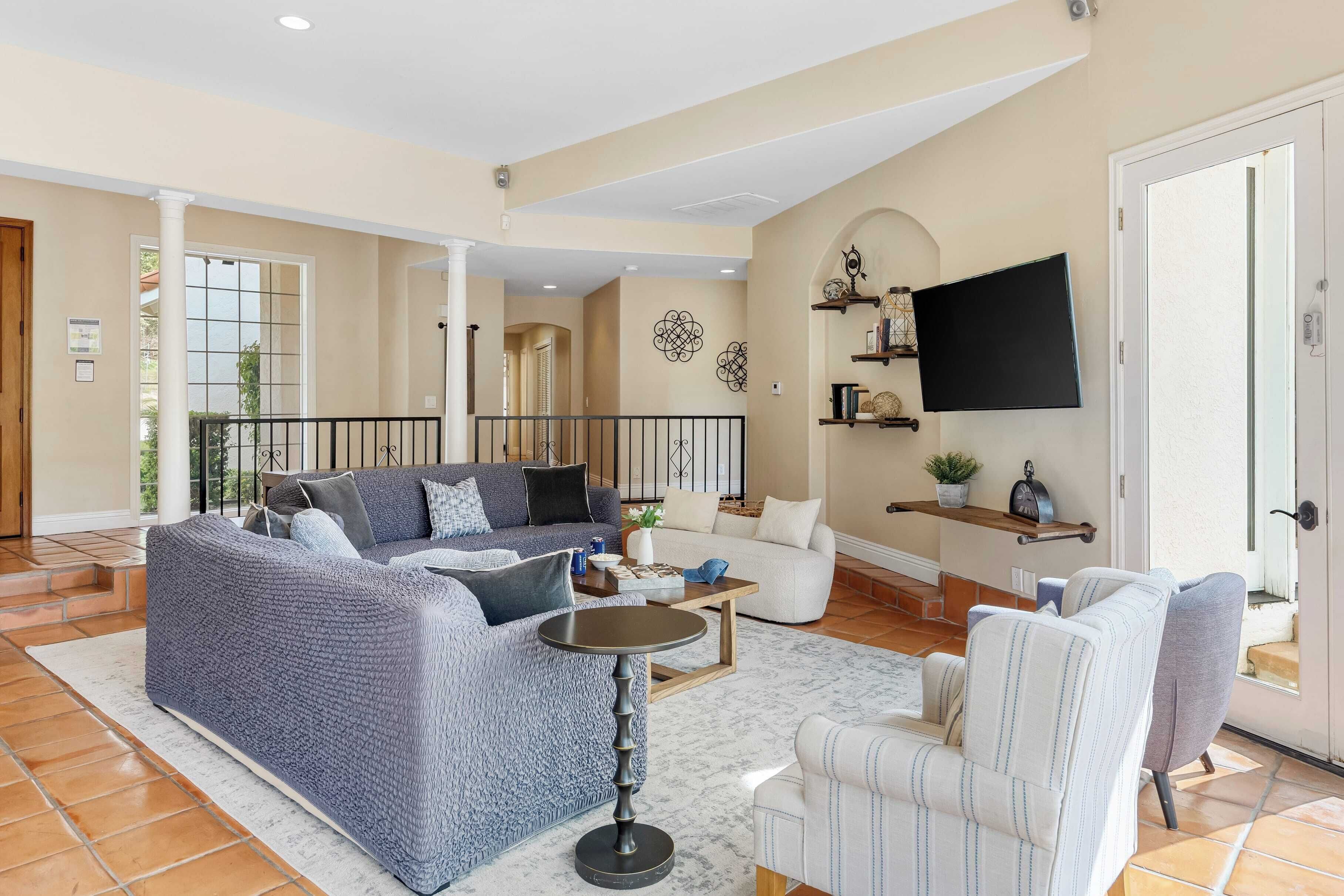 A spacious living room with a blue sofa, a white armchair, and a coffee table. There is a flat-screen TV mounted on the wall and a staircase leading to another level.