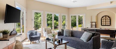 A spacious living room with a large sofa, a coffee table, and a flat-screen TV. There are large windows with a view of greenery outside.
