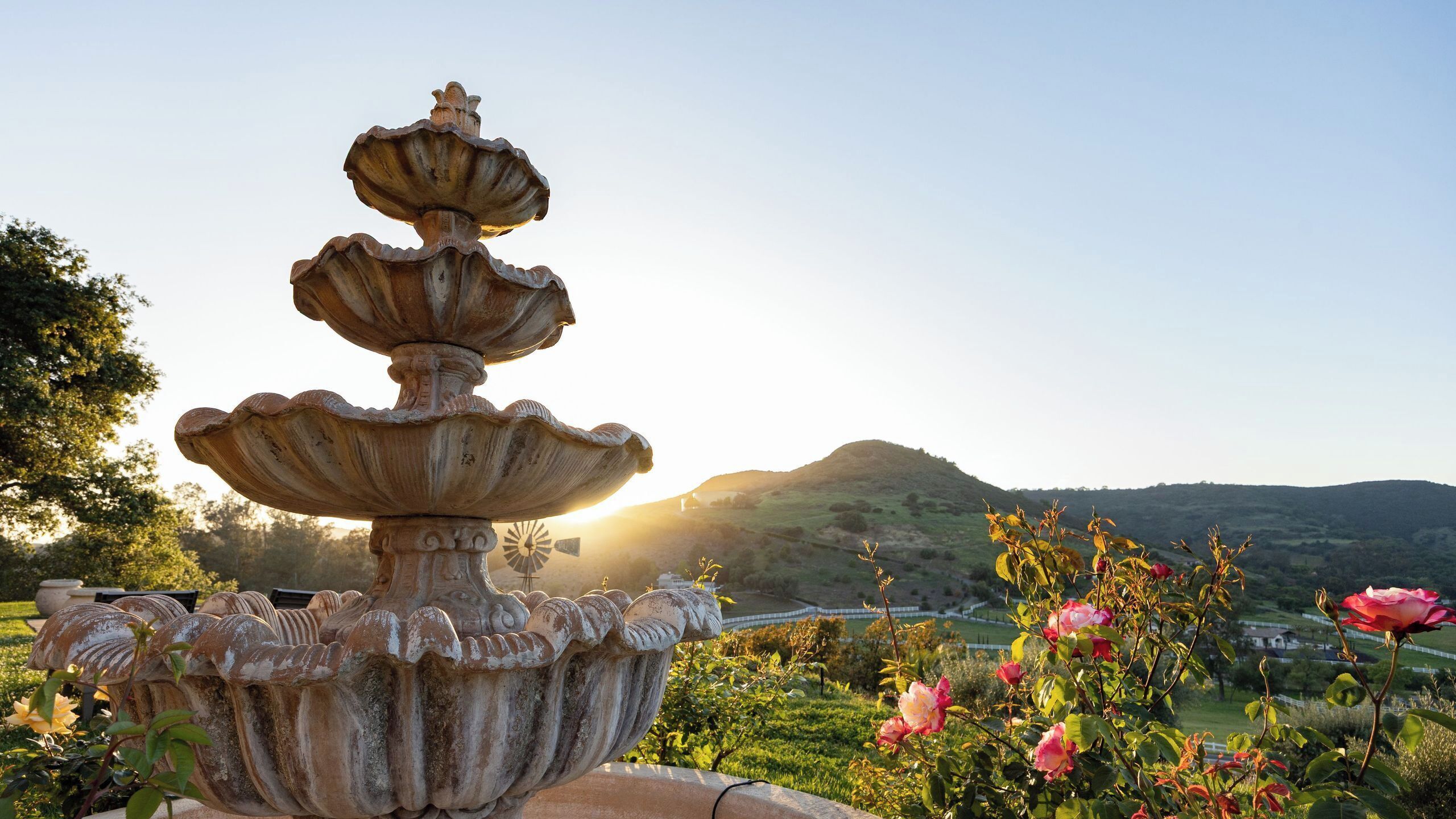 A multi-tiered stone fountain with intricate carvings, set in a garden with blooming roses and a backdrop of rolling hills.