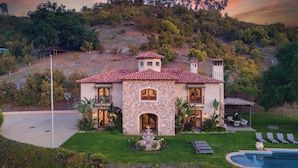 A two-story house with a stone facade, a red-tiled roof, and a swimming pool.