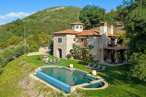 A large house with a swimming pool, a patio with lounge chairs, and a view of a hill.