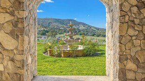 A stone archway framing a fountain with multiple tiers, set in a lush green landscape with a mountain in the background.
