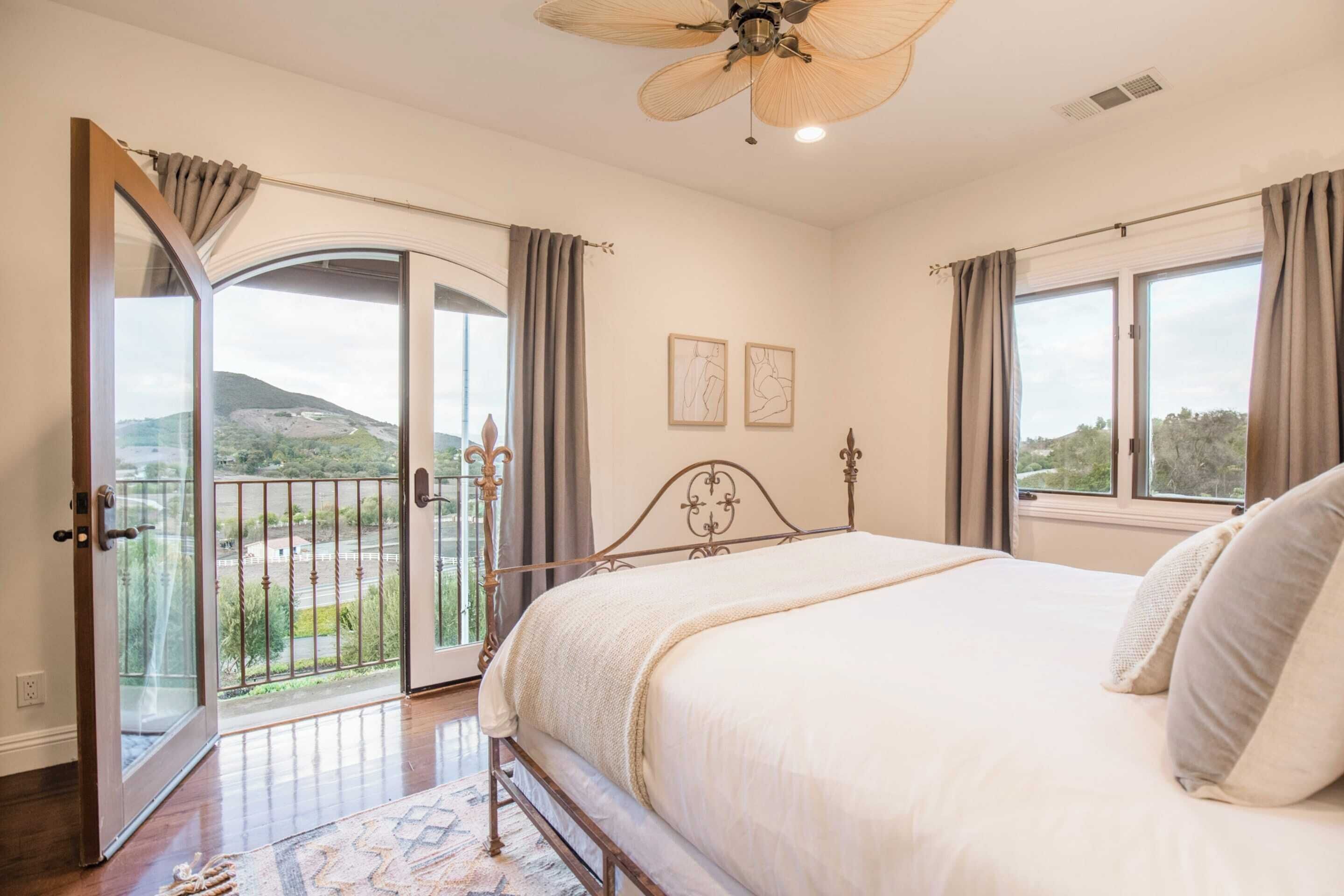 A bedroom with a large bed, a balcony with a view of the countryside, and a ceiling fan.
