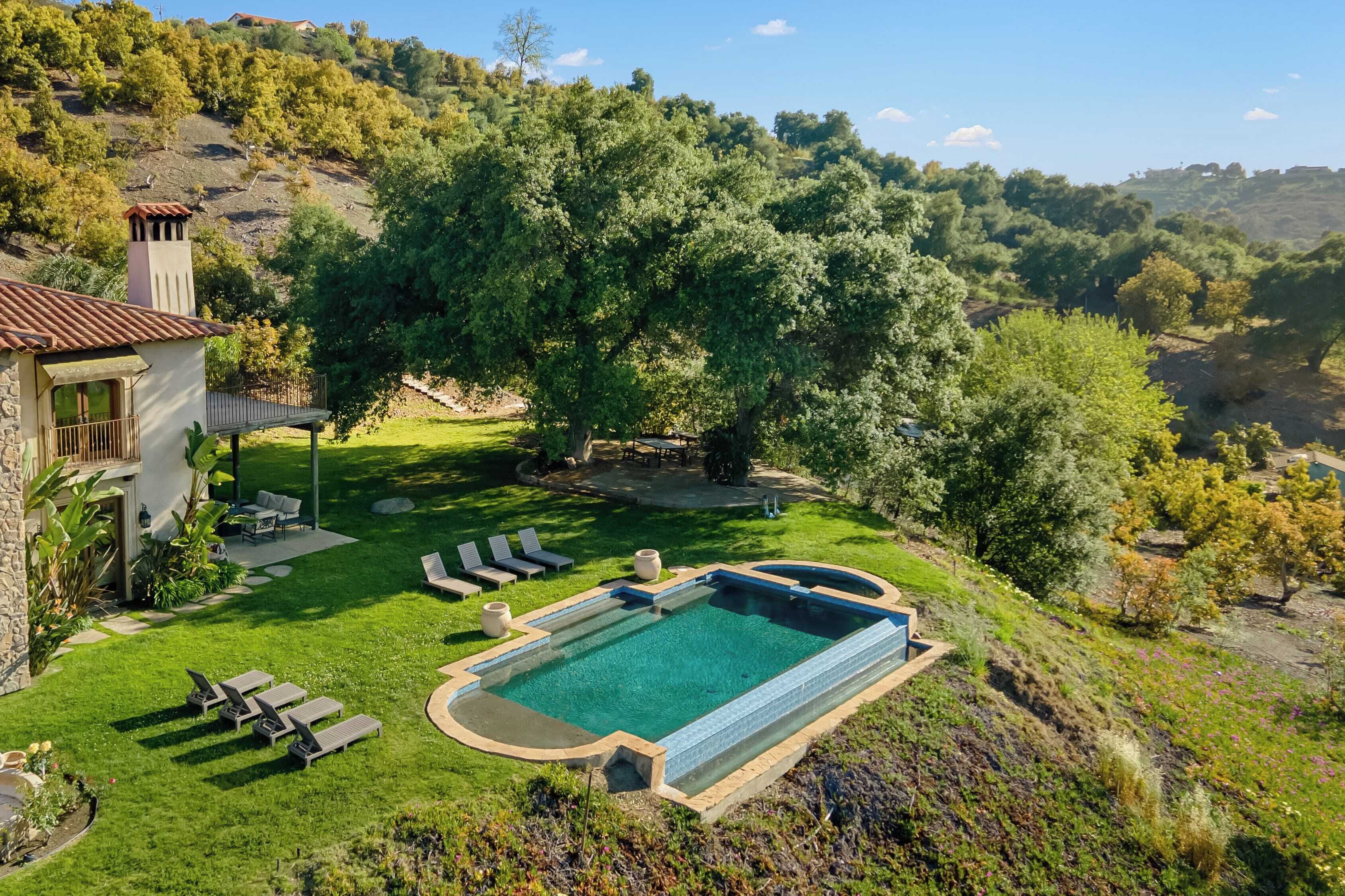 Une piscine entourée d’une pelouse bien entretenue, un bâtiment en pierre doté d’un balcon et une colline boisée.