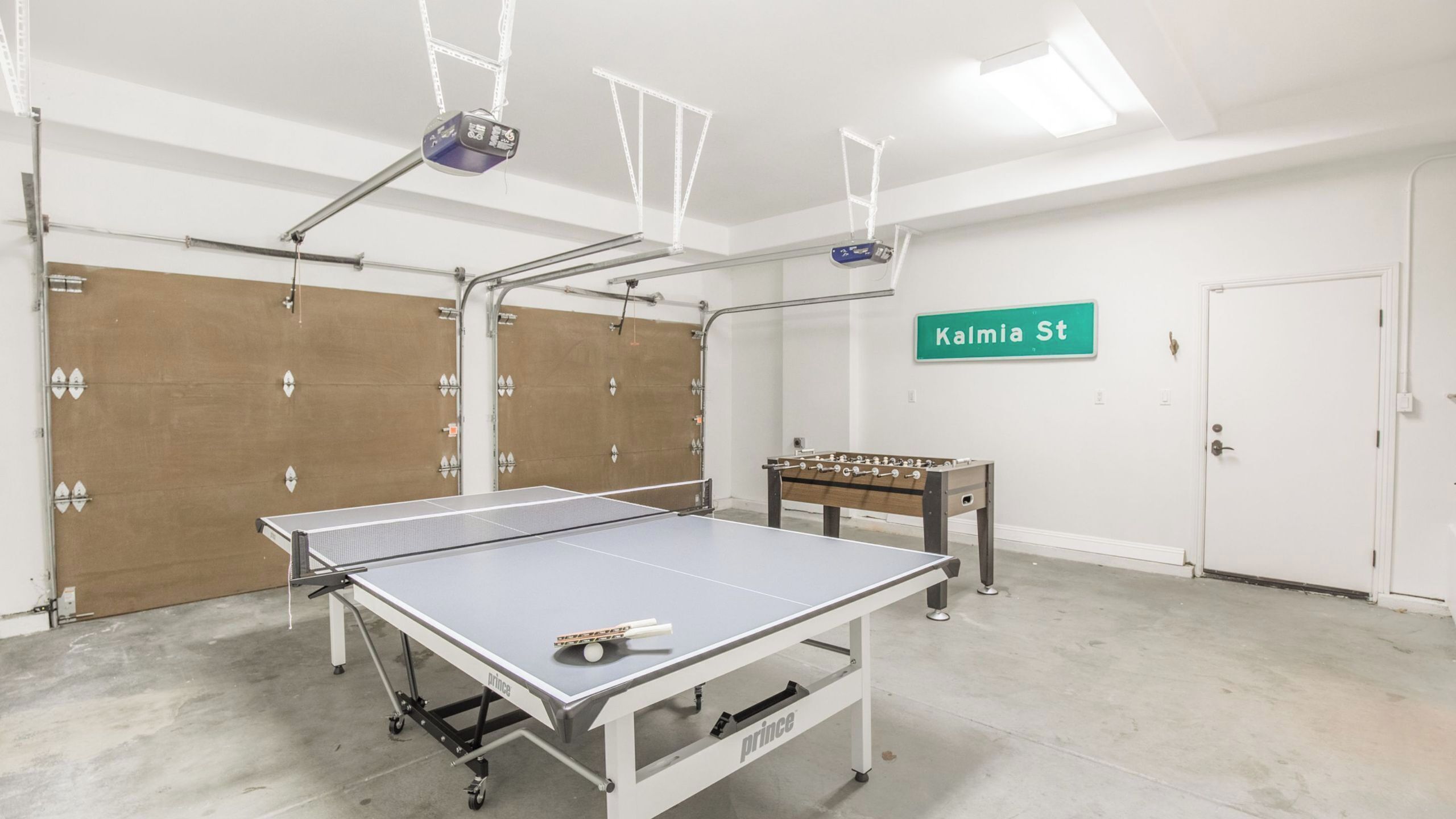 A ping pong table, a foosball table, and a green street sign reading 'Kalmia St' in a room with white walls and a concrete floor.