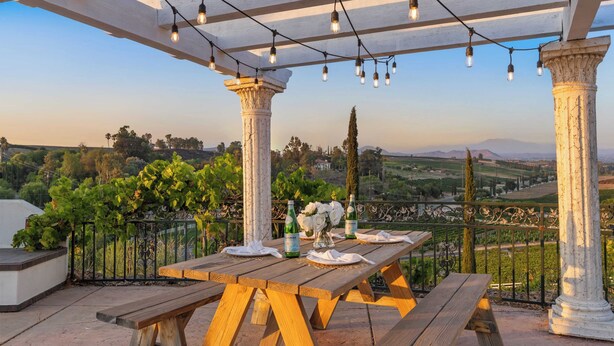 A wooden table set outdoors with a pergola overhead, columns, and a view of vineyards. - Wine Country Villa Large Rooftop Terrace w/ Views + Game Room Via del Sur (Temecula)