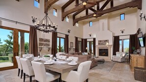 A spacious living area with a dining table set for six, a fireplace, and a large window overlooking a garden. - Owl's Nest by Avantstay Infinity Pool w/ Incredible Views (Temecula)