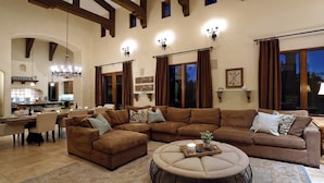 A spacious living room with a large sectional sofa, a round ottoman, and a dining area with a kitchen in the background. - Owl's Nest by Avantstay Infinity Pool w/ Incredible Views (Temecula)
