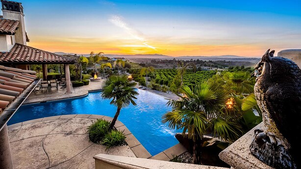 A swimming pool with a view of the sunset, surrounded by palm trees and a stone structure. - Owl's Nest by Avantstay Infinity Pool w/ Incredible Views (Temecula)