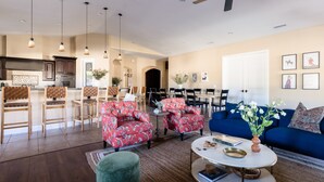 Amplia sala de estar con un sofá azul, sillones rojos con estampado floral y una mesa de centro redonda. Se ve una cocina con armarios de madera y un comedor con sillas al fondo.