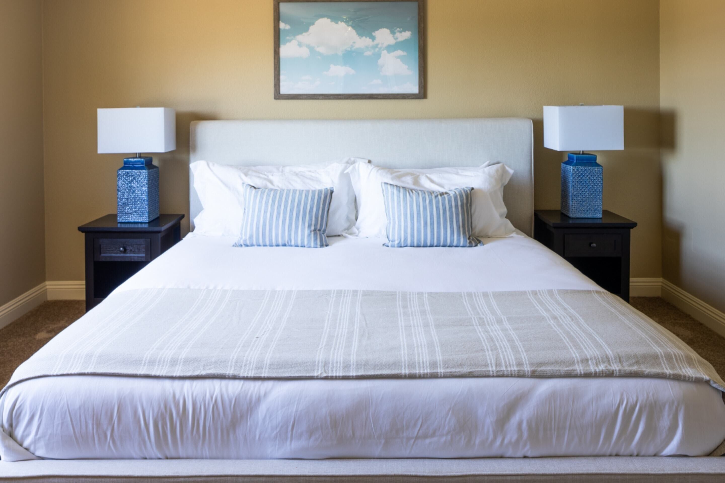A neatly made bed with white and striped pillows, two bedside tables with lamps, and a painting of clouds on the wall.