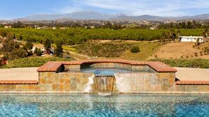 A swimming pool with a tiled hot tub and a waterfall feature, surrounded by a landscaped garden and a view of distant mountains. - Willow by Avantstay Mountain Views - See Hot Air Balloons From Pool (Temecula)