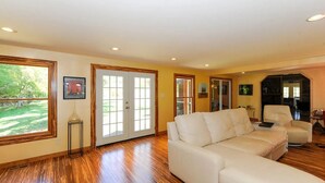 A spacious living room with a large white sectional sofa, wooden flooring, and large windows offering a view of the outdoors.