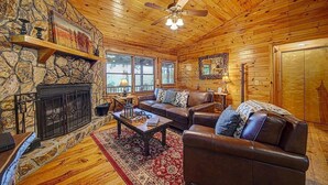 A cozy cabin interior with a stone fireplace, wooden walls, a leather sofa, and a ceiling fan. - Big Sky Lodge - stunning view (Blue Ridge)