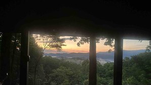 A view from a covered area with a wooden railing, overlooking a forested landscape with distant mountains and a sunset. - Big Sky Lodge - stunning view (Blue Ridge)