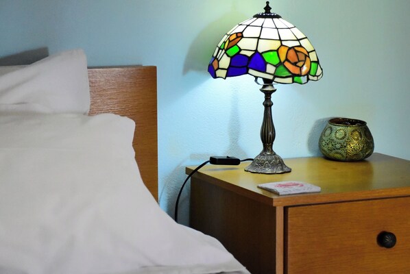 A bed with white sheets, a nightstand with a stained glass lamp, a power adapter, and a decorative vase.