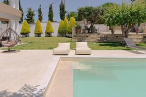 A swimming pool with two beige lounge chairs and a hanging chair, surrounded by neatly trimmed hedges and a stone wall. - Blue Sky Villa in Crete (heraklion)