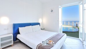 Une chambre avec un lit, une table de chevet, un plateau avec des tasses et des en-cas, et une vue sur la mer par une porte vitrée.