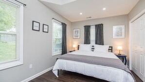 A bedroom with a large bed, two bedside tables with lamps, a window with blinds, and a door. - The Pink Pearl / 5 Min to Rupp / Newly Remodeled (Lexington)