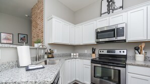 A modern kitchen with white cabinets, a granite countertop, stainless steel appliances, and a brick wall. - The Pink Pearl / 5 Min to Rupp / Newly Remodeled (Lexington)
