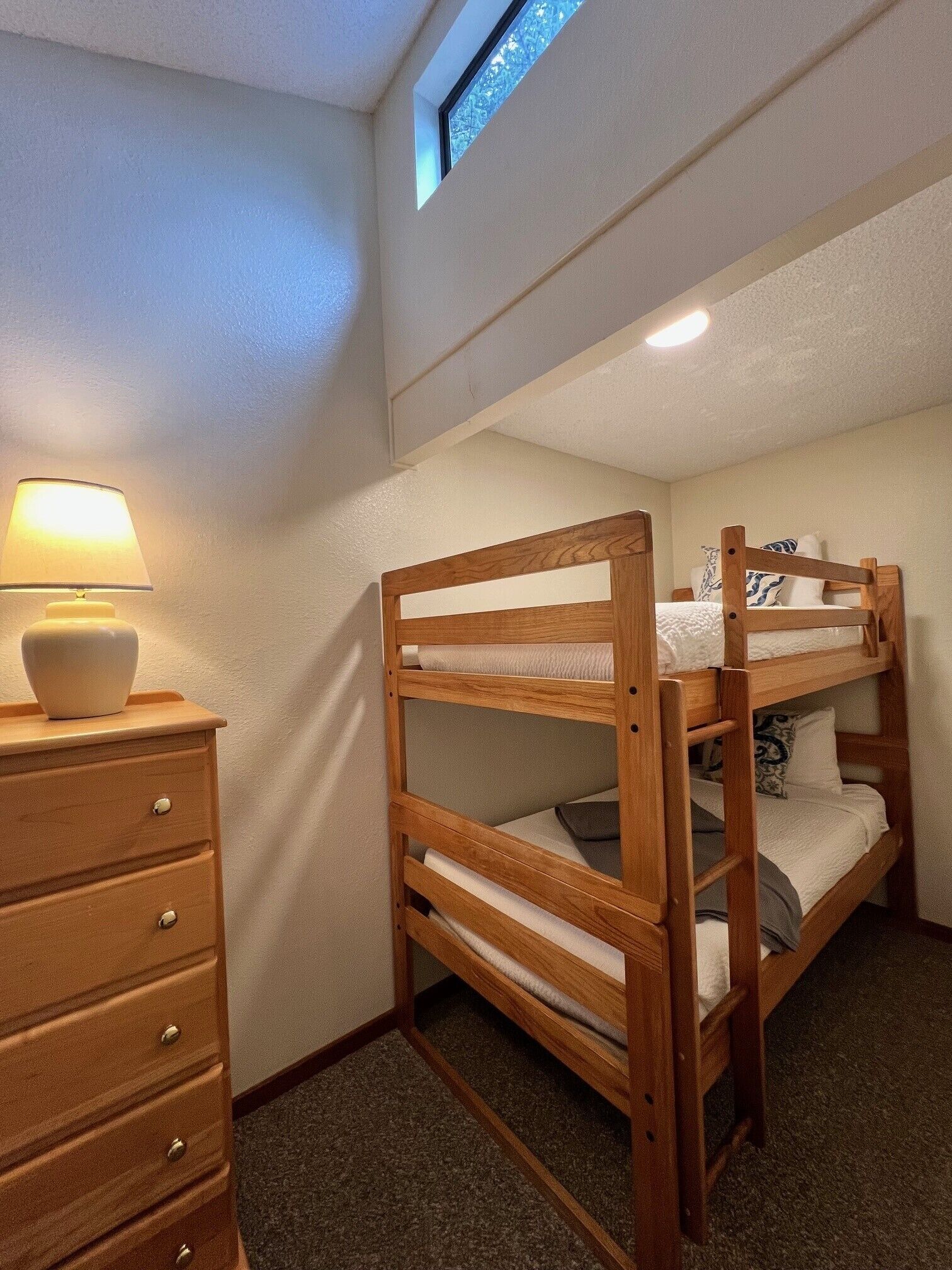 Una cama individual con estructura de madera, un aparador con cajones, una lámpara y un tragaluz.