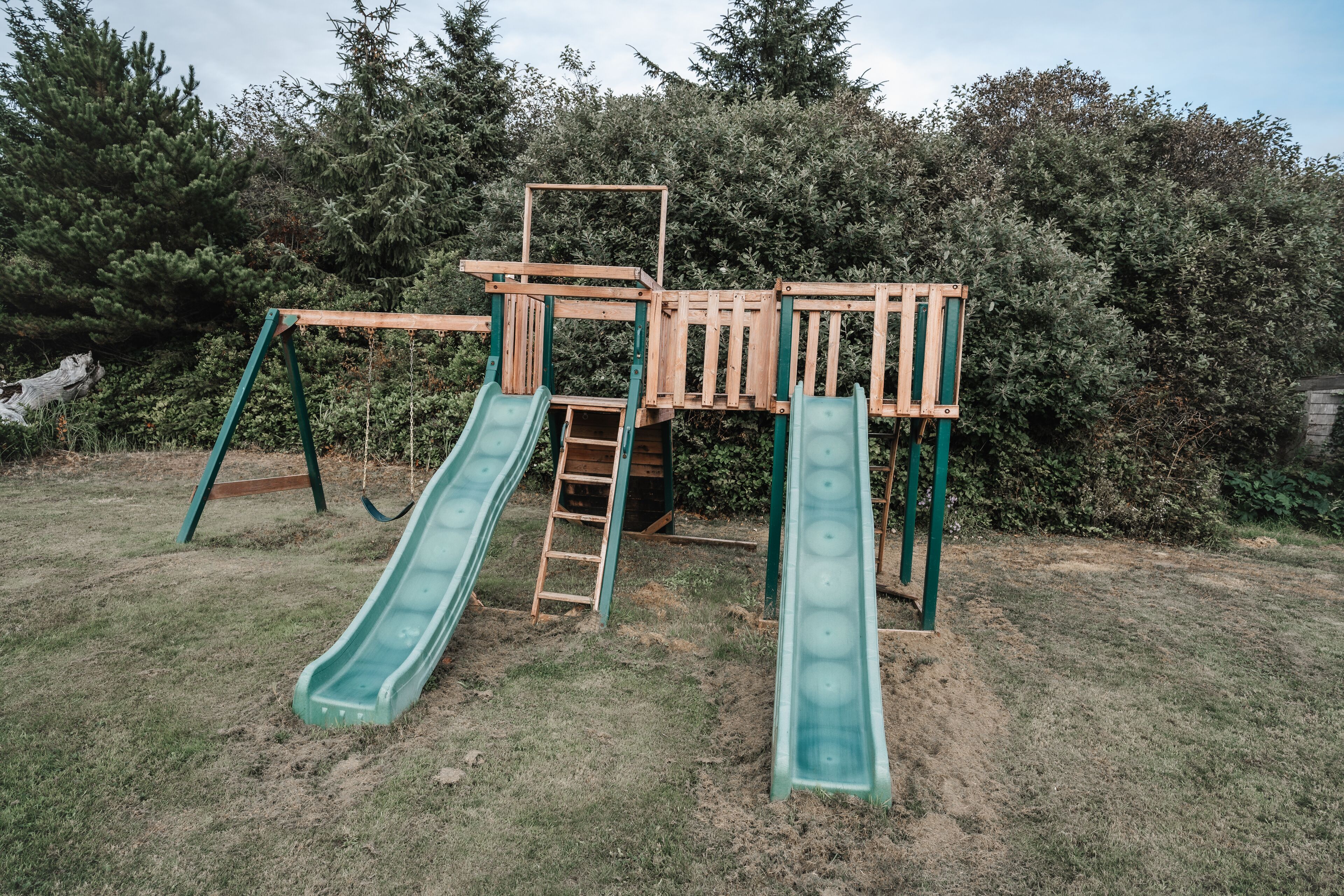 A wooden playground structure with two slides and a ladder, surrounded by dense foliage.
