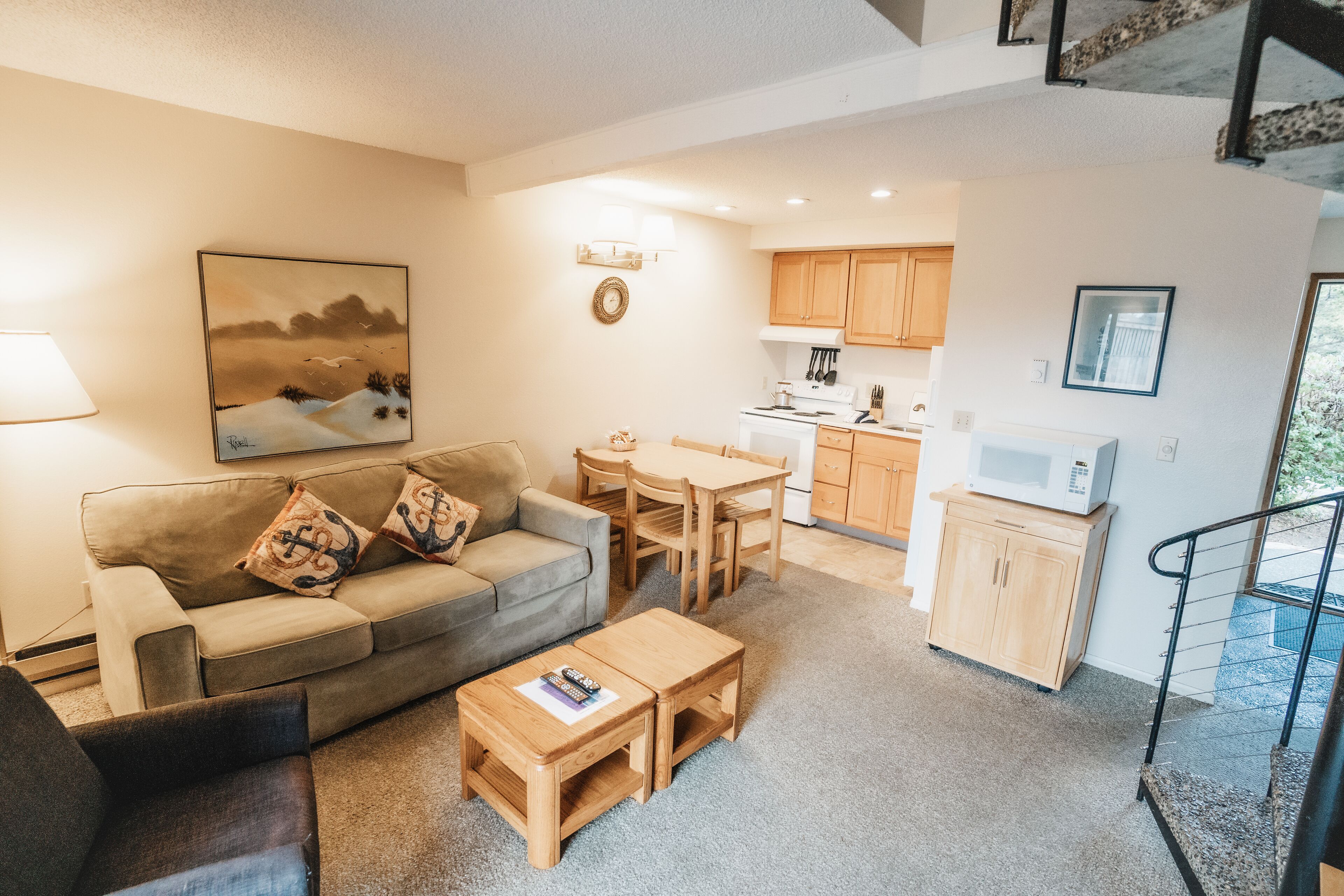 A cozy living room with a sofa, coffee table, and a painting. There is a dining area with a table and chairs, and a kitchen with cabinets and appliances.