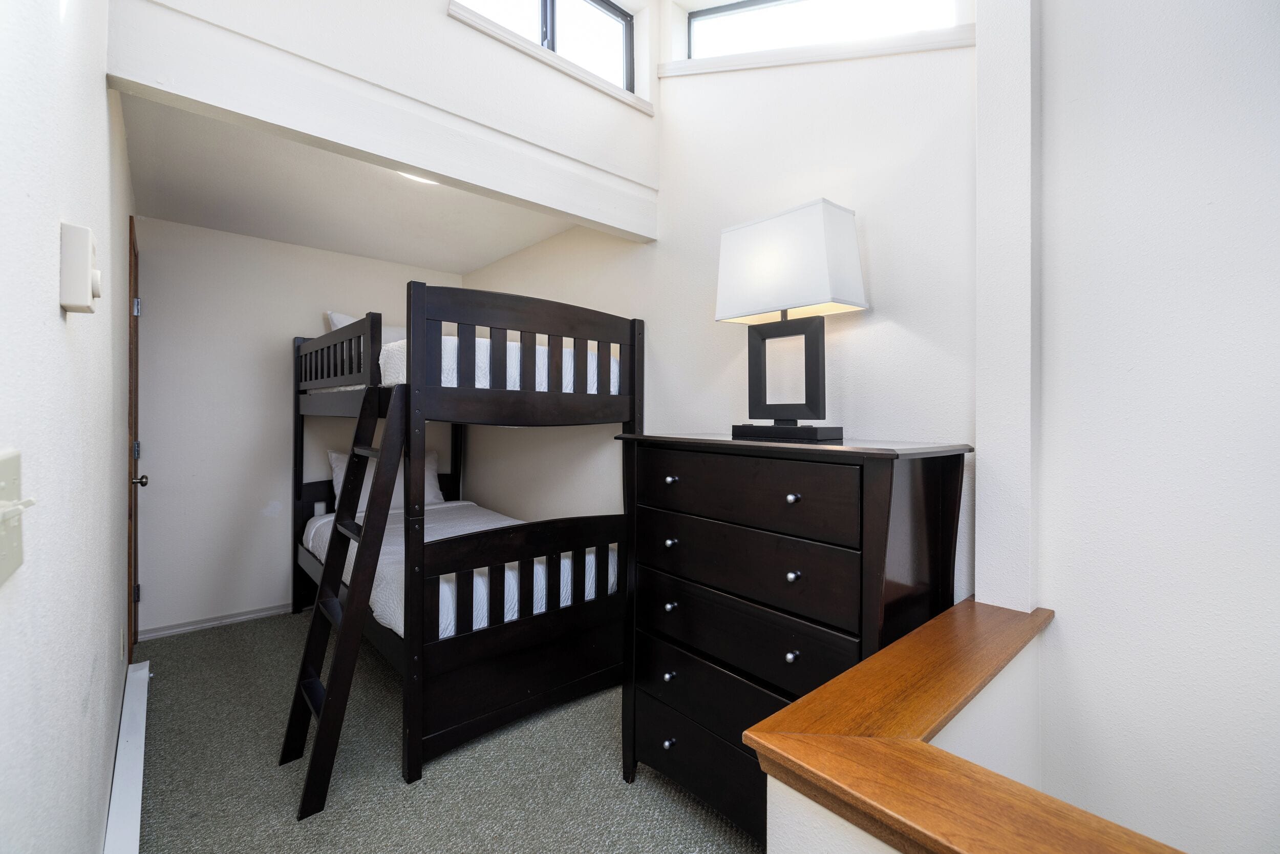 Una cama litera con escalera, un aparador con cajones, una lámpara y un banco de madera.