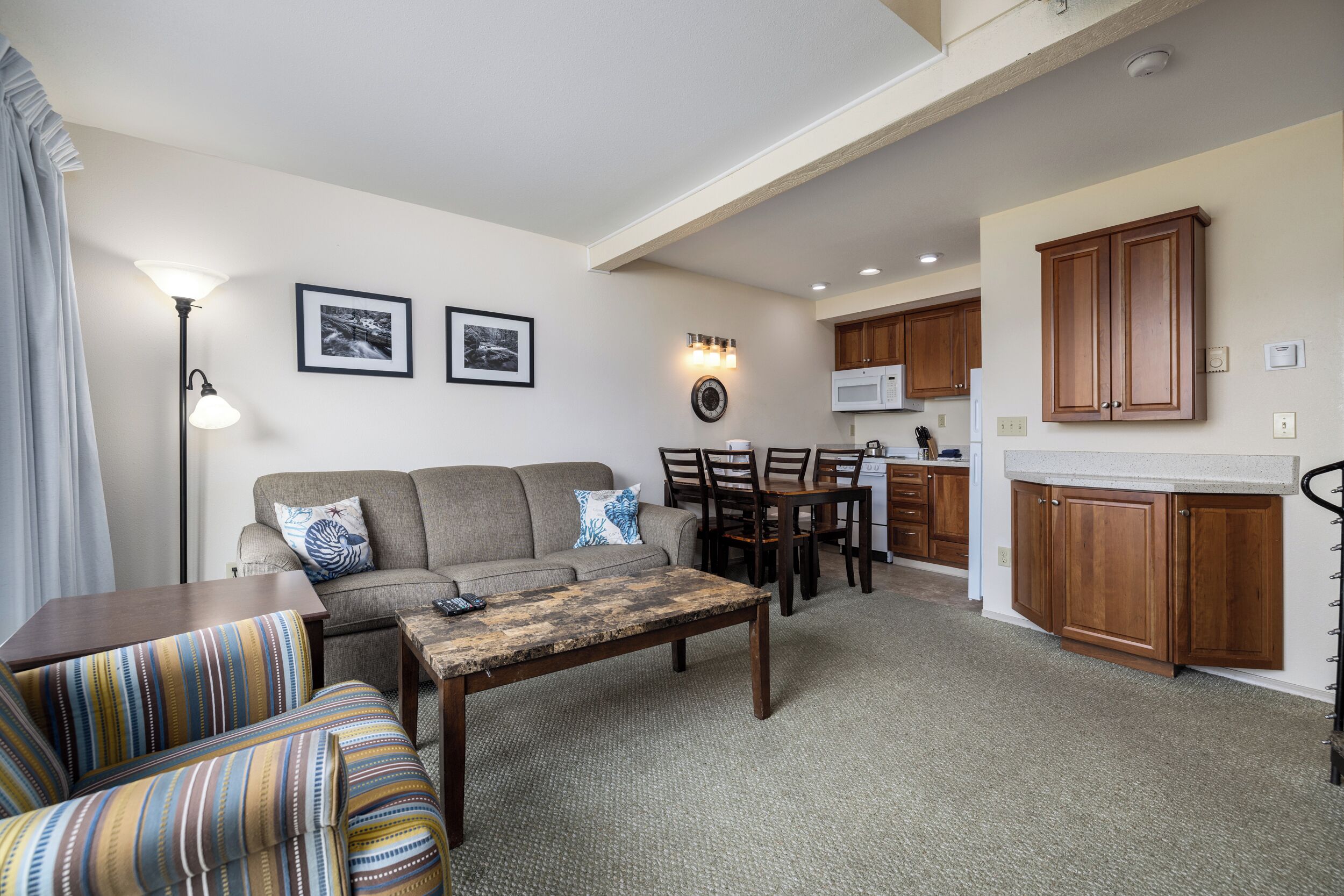 A living room with a sofa, a coffee table, and a dining area with chairs and a table. There is a kitchen area with wooden cabinets and a microwave.