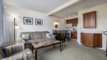 A living room with a sofa, a coffee table, and a dining area with chairs and a table. There is a kitchen area with wooden cabinets and a microwave.