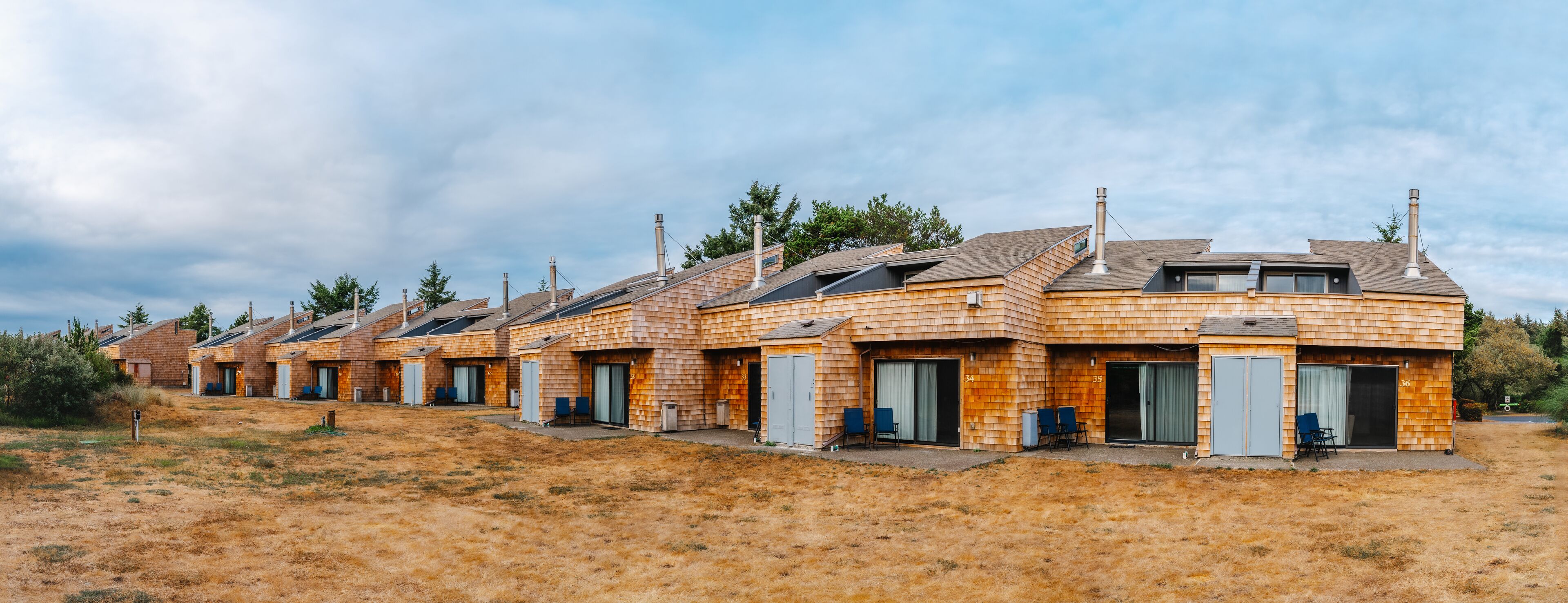 Een rij houten hutten met individuele deuren en ramen, genummerd op volgorde, gelegen in een droog, grasrijk gebied met bomen op de achtergrond.