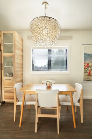 A dining area with a wooden table, white chairs, a decorative hanging light, a window with blinds, and a wooden cabinet. - Wallace BNB (Pinedale)