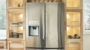A stainless steel refrigerator with a water dispenser, set in a kitchen with wooden cabinetry and a window with blinds.
