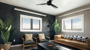 A living room with a dark wall, two windows with blinds, a ceiling fan, a brown leather sofa, a wooden coffee table, and a large green plant.