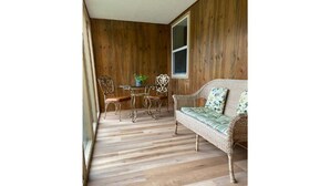 A room with a wooden floor, a wicker sofa with a floral cushion, a metal table with chairs, and a window. - The Farmhouse - Clean, comfortable, and affordable! (Fairbanks)