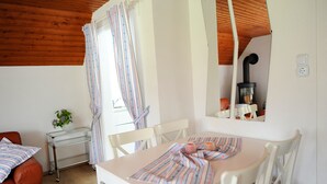 Una habitación acogedora con techo de madera, una mesa con sillas, una mesita blanca con una planta y una ventana con cortinas.