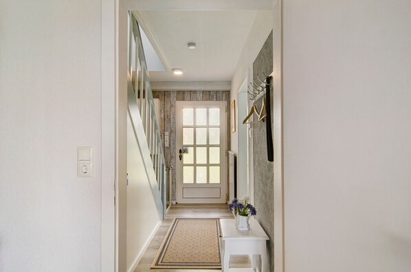 A hallway with a door, a wall-mounted coat rack, a small table with a vase, and a patterned rug. - Holiday Home 'Huus Karoline' with Garden & Wi-Fi (Hagermarsch Junkersrott)