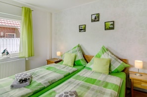 A double bed with green bedding, two bedside tables with lamps, a window with a curtain, and three framed pictures on the wall.
