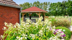Ein Backsteinhaus mit rotem Dach, einem hölzernen Pavillon und einem gepflegten Garten mit verschiedenen Pflanzen und Blumen.