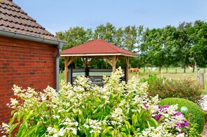 A brick building with a red roof, a wooden gazebo, and a well-maintained garden with various plants and flowers. - Holiday Home 'Huus Karoline' with Garden & Wi-Fi (Hagermarsch Junkersrott)
