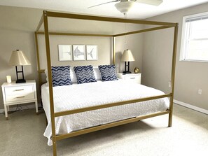 A four-poster bed with a gold frame, white bedding, and patterned pillows. There are two bedside tables with lamps and a wall-mounted light fixture.