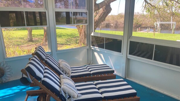 Une véranda avec un fauteuil de relaxation en bois, des coussins à rayures et une vue sur une zone herbeuse et un plan d’eau.