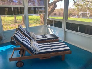 Une véranda avec un fauteuil de relaxation en bois, des coussins à rayures et une vue sur une zone herbeuse et un plan d’eau.