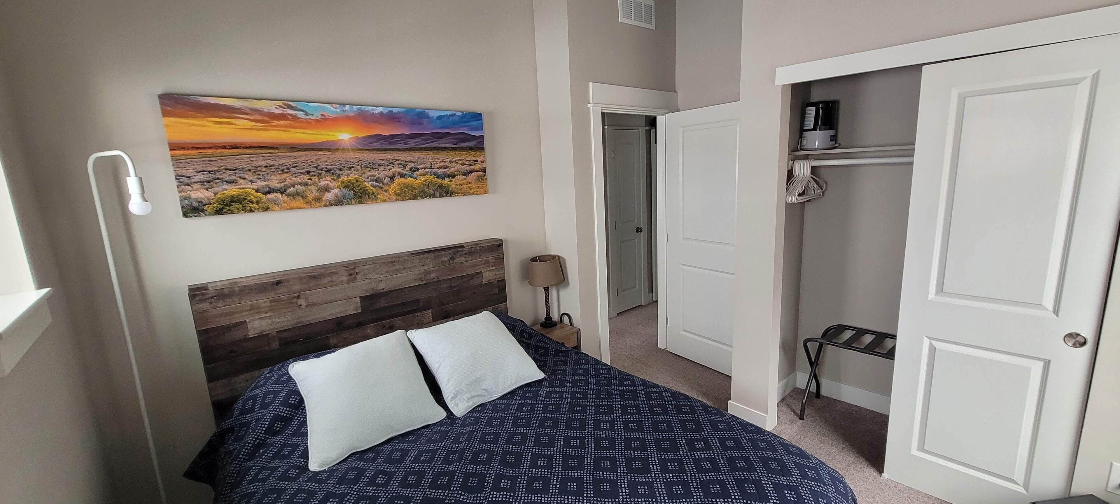 Une chambre à coucher avec un lit, une table de chevet, une lampe, un tableau représentant un coucher de soleil et une armoire dont la porte est ouverte.