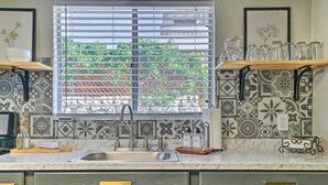 Cocina con lavamanos, ventana con persianas, salpicadero estampado y estantes con cristalería.
