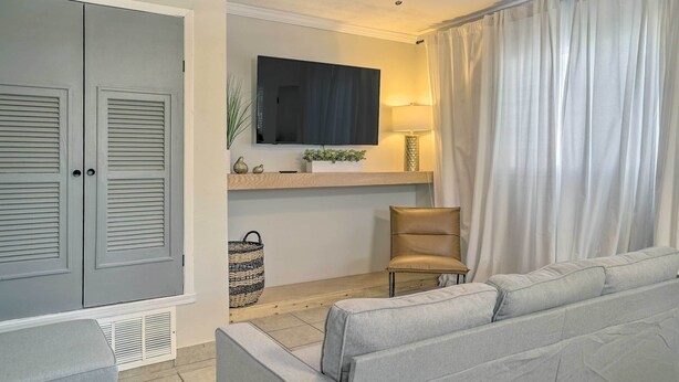 Una sala de estar con un sofá gris, un sillón color crema, un televisor de pantalla plana en la pared, un ventilador de techo y un ventanal con cortinas blancas.